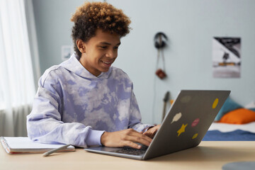 Smiling diligent teenager in hoodie typing on laptop keyboard while sitting by desk in home environment and preparing homework - Powered by Adobe