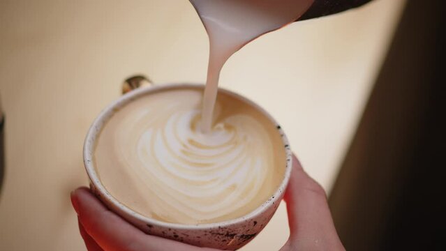 Pouring foamed milk into a beautiful cup of coffee. The barista prepares a delicious cappuccino at the bar. Pleasant warm atmosphere of the cafe