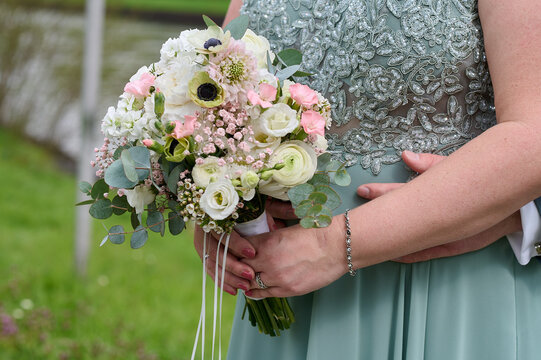 Frau in einem lindgrünen Kleid hält einen pastell farbenen Brautstrauß vor dem Bauch, Hand des Partners unterstützt von hinten