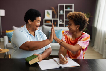 Happy teenage schoolboy and mature female tutor giving each other high five while sitting by table...