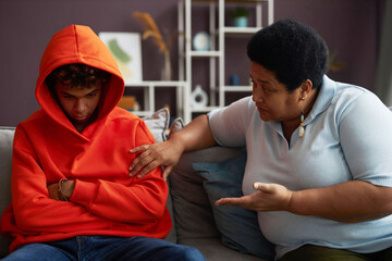 African American mature woman talking to sulky or stressed teenage grandson in red hoodie while...