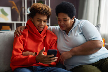 African American teenage boy in red hoodie showing online video in smartphone to his grandmother while both sitting on couch