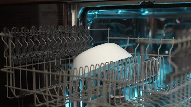 Close-up hands of unrecognizable man loading backlit dishwasher machine with white deep plates. Concept using modern house kitchen appliance to keep home clean. Shooting in slow motion.