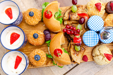 Breakfast eating concept with various morning food placed on a wooden plate: yogurt with fruits salad, sandwiches, crepes, jams, cookies and cupcakes.