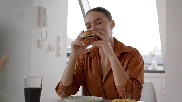 Young woman dining at home, eating fresh burger and french fries with pleasure, tracking shot, slow motion, free space