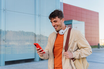 Cheerful young man with smartphone on street. Low angle of young male with headphones on neck browsing mobile phone while standing on city street and looking at camera
