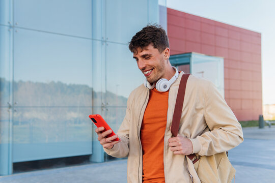 Cheerful young man with smartphone on street. Low angle of young male with headphones on neck browsing mobile phone while standing on city street and looking at camera