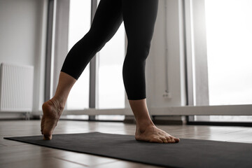 Close up view of athletic female in tight pants standing barefoot on black mat while getting into yoga position in bright fitness studio. Sporty woman performing daily workout practice indoors.