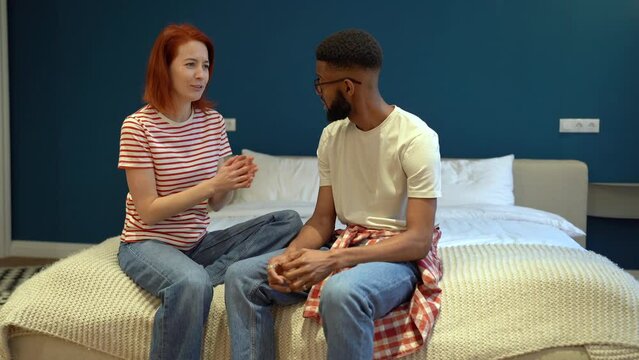 Loving couple sitting in bed after work day laughing gesturing share good news each other. Happy smiling european woman and african american man chatting, talking, tell funny stories, interested ideas