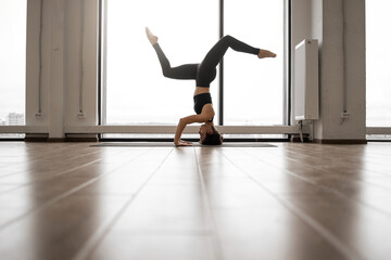Relaxed dark haired woman using yoga mat for doing Pincha Mayurasana exercise at spacious fitness studio. Caucasian female in sport attire training regularly for body care.