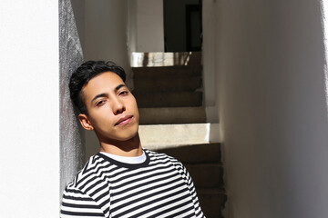 Handsome latin guy enjoying the moment and basking in the sunlight. Portrait of young man of hispanic heritage wearing striped shirt leaning against the white wall.