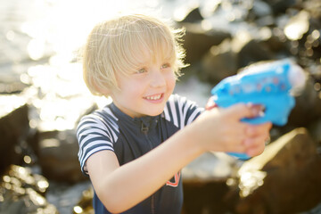 Fototapeta premium Cute little boy playing with water pistol on the seashore on a hot day. The child having fun with water gun outdoors during the summer holidays.