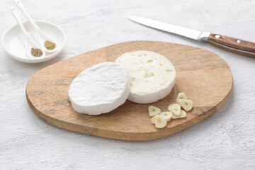 Wooden board with a cut head of white mold cheese, chopped garlic and spices on a light gray background. Stage of cooking a delicious holiday snack