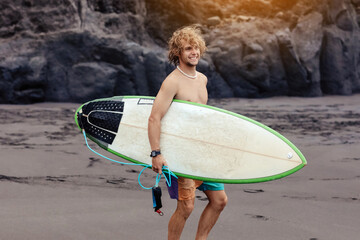 Fit young surfer man with curly blond hair with surfboard goes by the ocean having fun doing extreme water sports, surfing. Travel and healthy lifestyle concept. Sports travel destination.