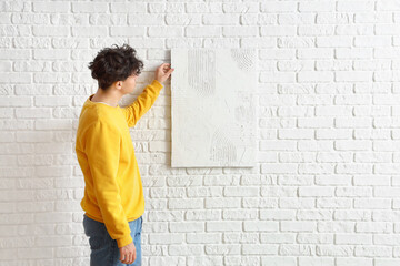 Young man hanging painting on white brick wall