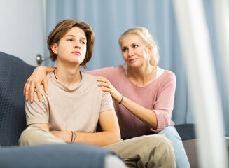Obraz premium Portrait of upset and offended teenager sitting on sofa while mother soothing him. Mood swings and puberty concept