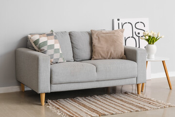 Cozy sofa with cushions and tulip flowers in vase on table near grey wall