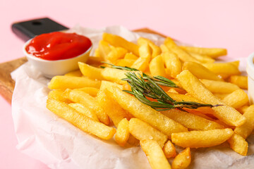 Wooden board with tasty french fries, rosemary and ketchup on pink background, closeup