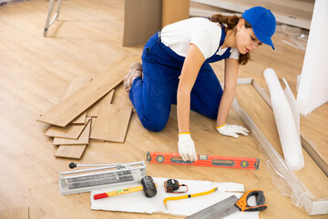 Young woman builder installing parquet in construction site, measuring floor with level.