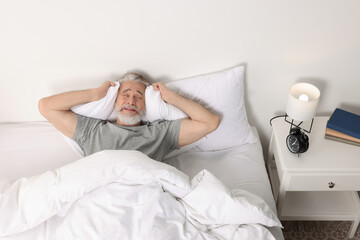 Senior man covering ears with pillows in bed and alarm clock on bedside table at home