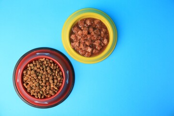 Wet and dry pet food in feeding bowls on light blue background, flat lay. Space for text
