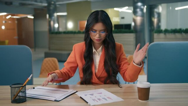 Tired stressed girl looking at reports tables, grabs her head with hand while working hard at late night at empty office. Exhausted asian business woman reviewing documents on desk with paperwork 4K