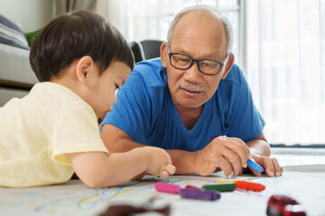 Asian Senior man and little child boy lying on floor drawing and coloring with Crayons together. Grandfather and Grandson painting with colorful Crayons on paper.