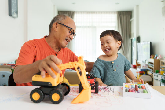 Happy Asian Grandfather and Grandchild laughing while drawing and coloring with Crayons together. Playful Chinese Senior man and little child boy painting on paper.