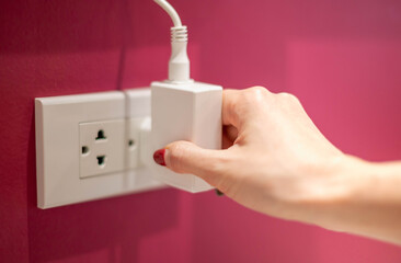 The hand is unplugging the electric line off a white socket on the red wall.