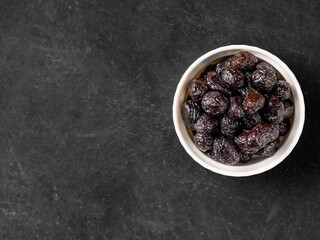 Black olives in white bowl on black background. Dried ripe fruits of Olea europaea, one of core ingredients in Mediterranean cuisine. Edible, raw, organic food. Top view, copy space, close up.