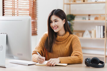 Online education, e-learning. Asian woman in stylish casual clothes, studying using a computer, listening to online lecture, taking notes, online study at home