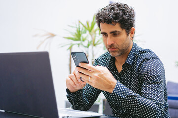 Middle-aged Latin man in shirt interacting with his cell phone in front of his laptop at his workplace. Copy space.