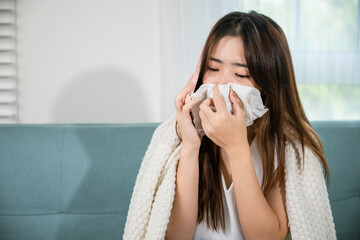 Asian young woman sick sitting down on sofa with cold blowing her nose hold tissues and talking on cellphone to doctor employer while asking about sick leave with mobile phone in living room at home