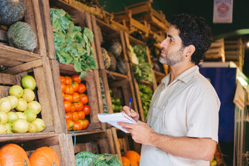 Middle-aged Latin man working in a grocery market while making notes in front of some crates used as shelves.