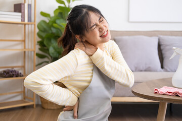 Asian woman doing housework wearing apron sitting on floors with shoulder feeling hurt painful in the living room. young lady Cleaning table by spraying Cleaning Products and wiping..