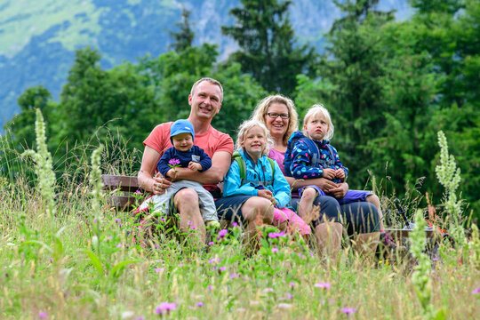 Fröhliche, junge Familie posiert auf einer Bank inmitten einer Wiese im Gebirge