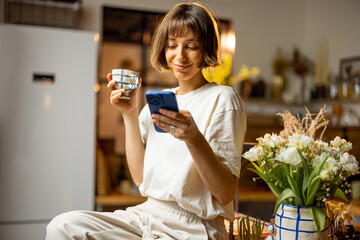 Young woman uses smart phone and drinks a coffee, spending morning time on beautiful kitchen with...