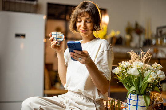 Young woman uses smart phone and drinks a coffee, spending morning time on beautiful kitchen with fresh flowers at home