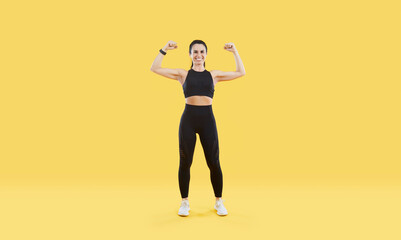 Fototapeta na wymiar Fit and healthy woman. Positive sporty woman showing biceps and strong arms isolated on vivid yellow background. Portrait of woman with slender figure dressed in black sportswear set. Full length.