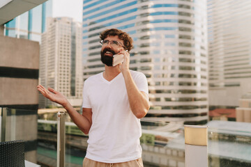 Handsome man with beard  talking by mobiyle phone while walking in big modern city with skyscrapers. Outdoors at sunset . Wearing white t shirt.