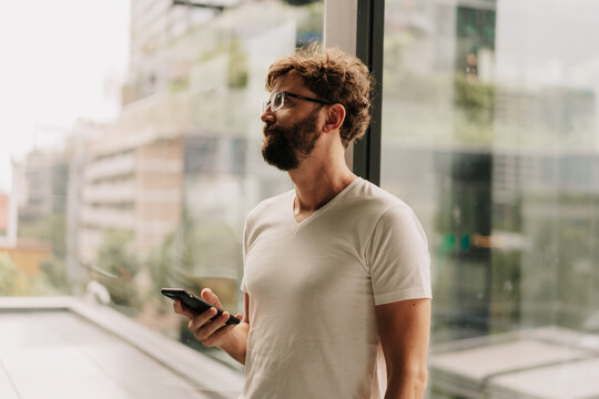Handsome man with beard using mobiyle phone while walking in big modern city with skyscrapers. Wearing white t shirt.