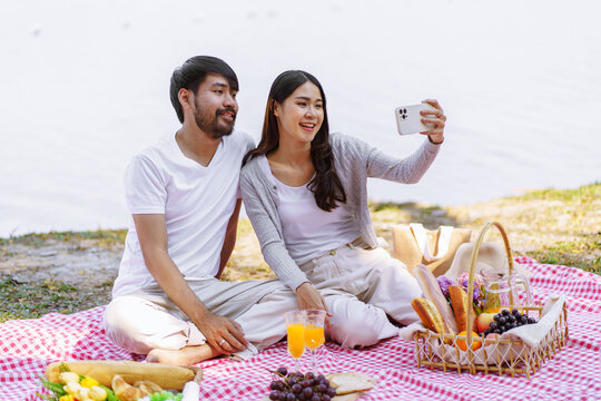  In love asian couple taking selfie photo with smartphone relaxing together with picnic Basket