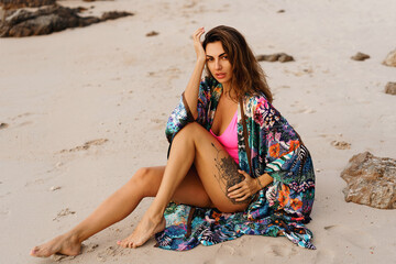 Geourgeous sensual  woman in pink swimsuit, and boho kimono  with tropical print  posing on  the beach at sunset.