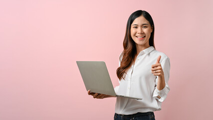 Image of young Asian woman holding laptop and give a thumbs up sign, pink isolated background.