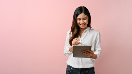 Young beautiful Asian woman writing on digital tablet, pink isolated background.