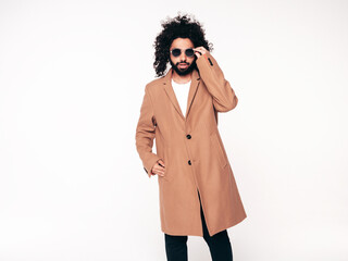 Handsome smiling hipster model.  Unshaven Arabian man dressed in brown coat clothes. Fashion male with long curly hairstyle isolated on white in studio. Cheerful and happy. In sunglasses