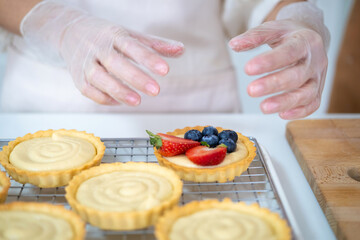 Obraz na płótnie Canvas close up of hands in cooking gloves Baker adding blueberries strawberry fresh fruit to a tart on white table in Kitchen. housewife baker wear apron making fruit tart. homemade bakery at home.