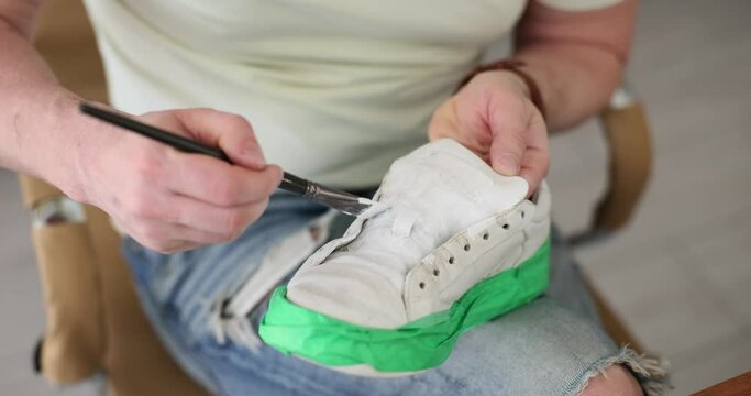 Man paints leather sneakers with white paint in workshop. Sports shoe repair