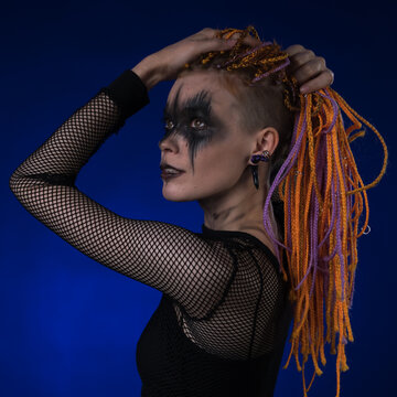 Dramatic portrait of young female with spooky black stage makeup painted on face and orange color dreadlocks hairstyle. Side view, studio shot on blue background. Part of photo series