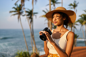 Young african female model tourist with camera in colorful clothes and straw hat taking pictures at tropical location at sunrise. Black travelling woman takes photos in exotic ocean scenery at dawn. - Powered by Adobe
